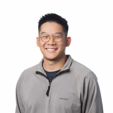 Asian man with glasses smiling, casual grey jacket, friendly expression.