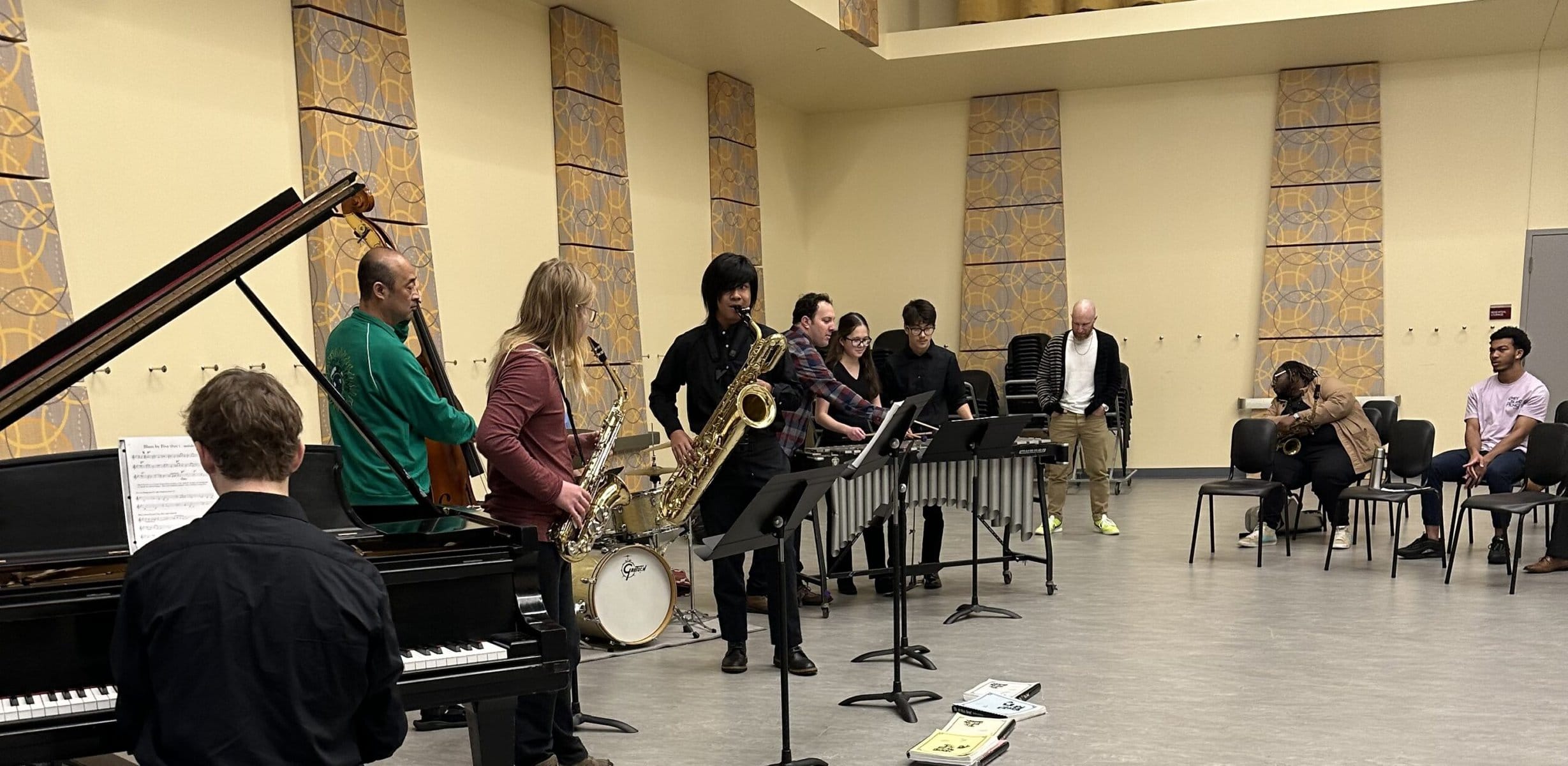 Jazz ensemble performing live at Madison Jazz Society event in a spacious room with acoustic panels. Musicians include saxophonists, pianist, bassist, and percussionists, showcasing jazz music excellence.