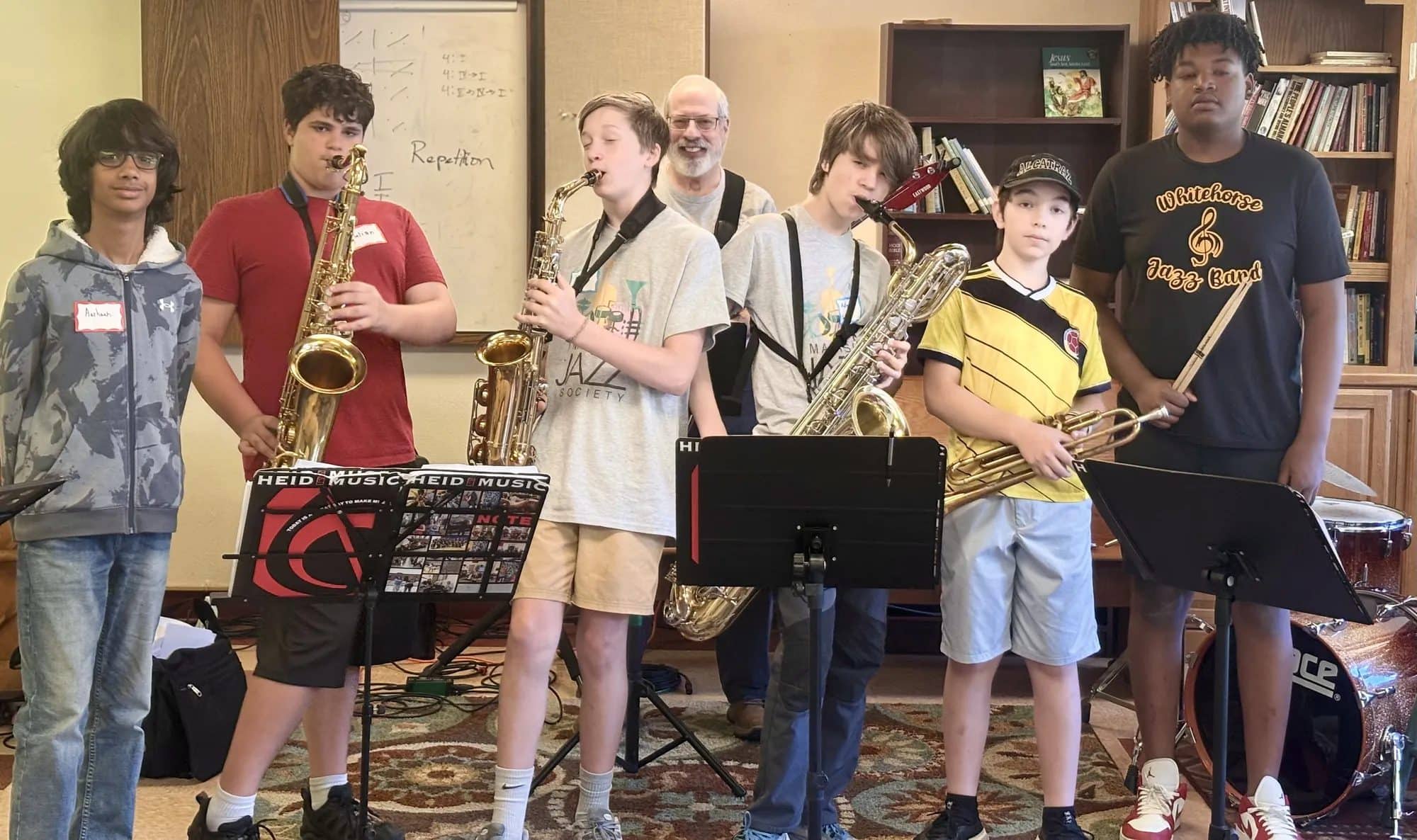 Young musicians with saxophones, a baritone horn and drumsticks stand together during an instrument-specific session at iGnitE jAzz!, with an instructor behind them