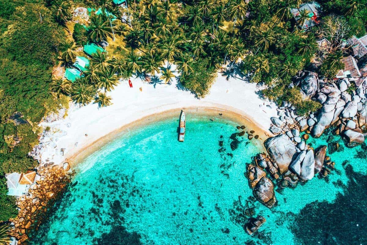 An aerial view of a tropical cove in Thailand with white sands, palm trees and a traditional boat