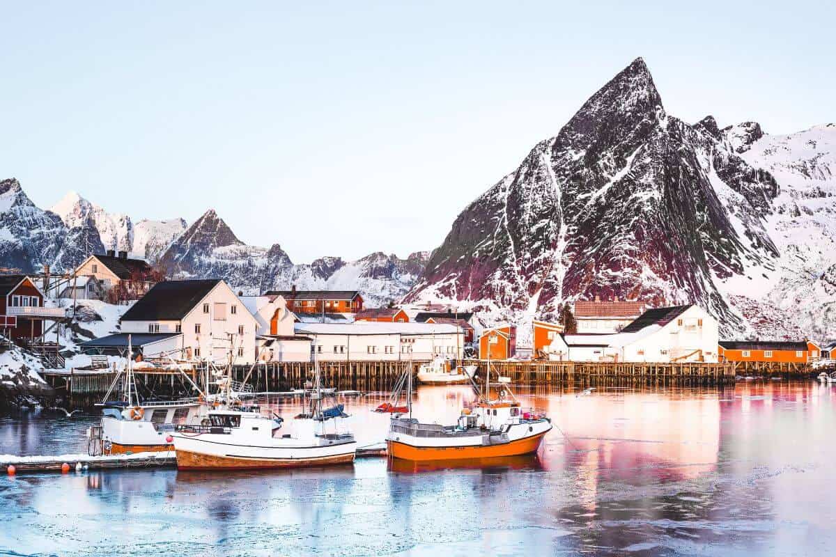 A quaint, snowy harbour in Norway with red boats and buildings in front of rugged mountain scenery