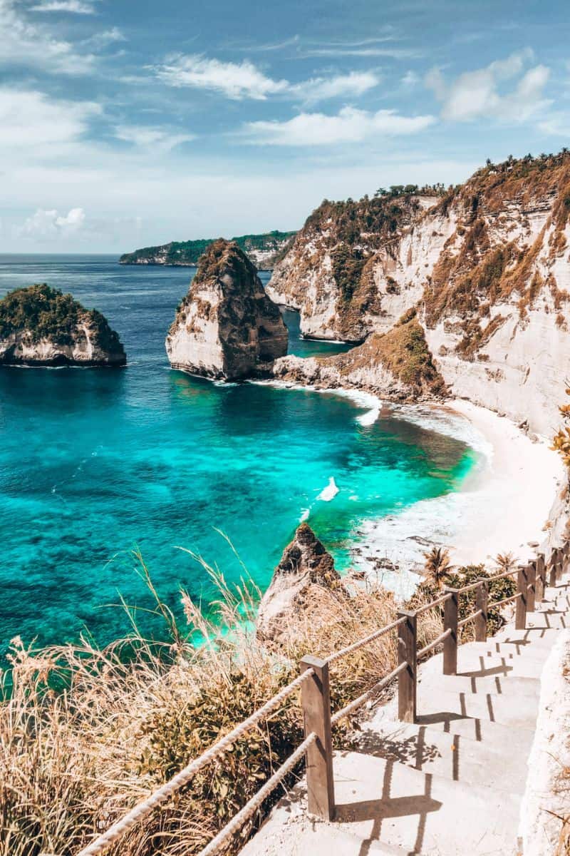 A staircase along the side of a rugged cliff heading towards a tropical white-sand beach in Nusa Penida, Indonesia