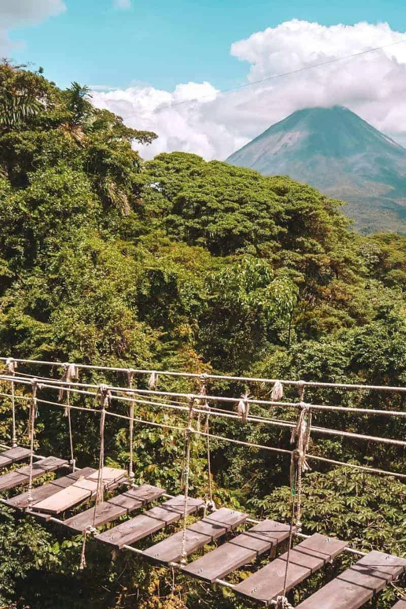 A wooden suspension bridge across the rainforest in Costa Rica with a volcano in the background