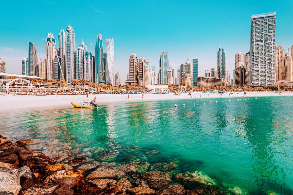A view across water towards a sweeping white-sand beach, backed by skyscrapers in Dubai