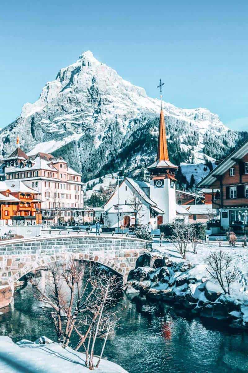 A picturesque Swiss town with a church spire and traditional buildings in front of a snowy mountain and icy river 
