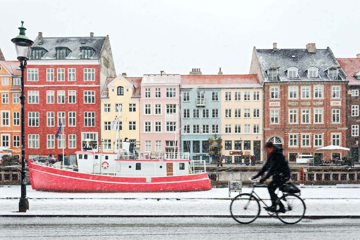 A man riding a bike in front of a snowy canal with a red boat in Copenhagen's Nyhavn waterfront district