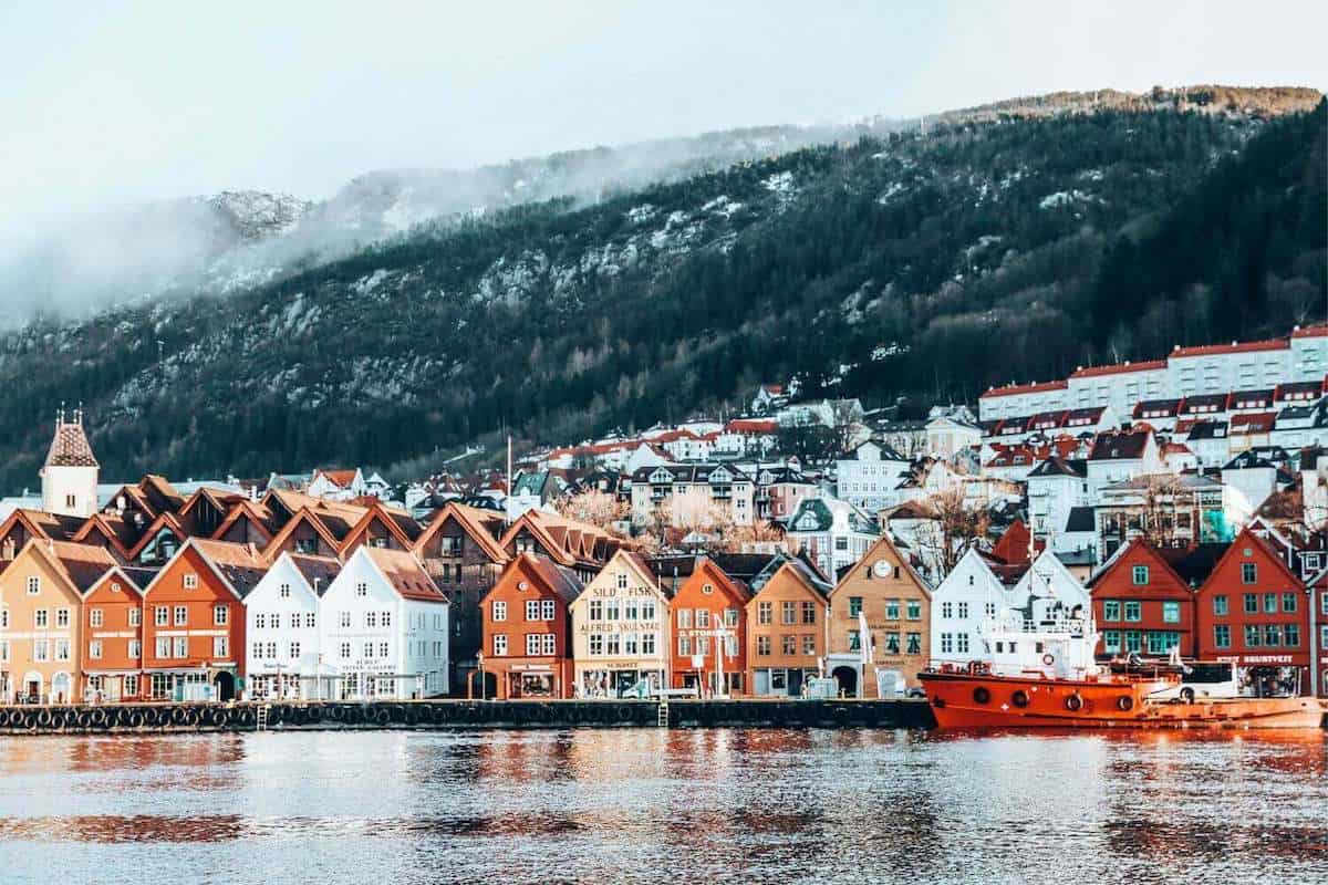 A view across an empty harbour towards colourful orange and red houses under snowy mountains in Bergen, Norway