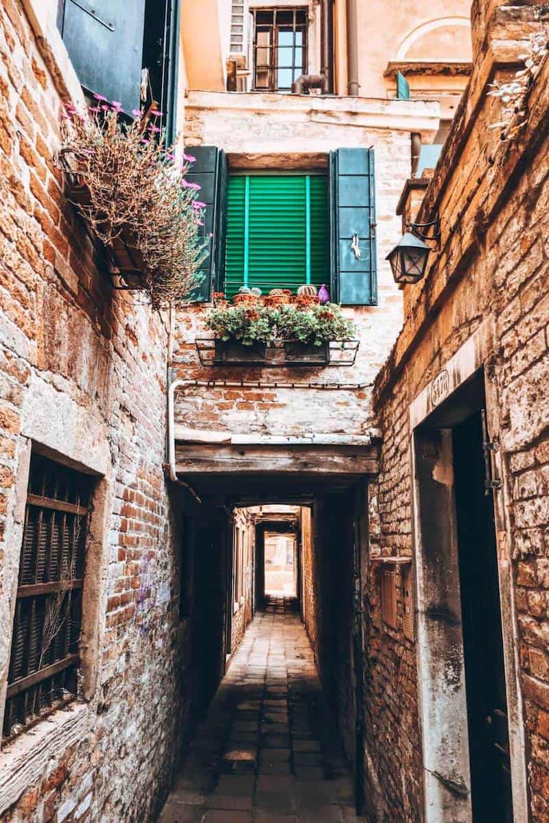 A quaint and narrow alleyway in Venice going under a building with blue shutters