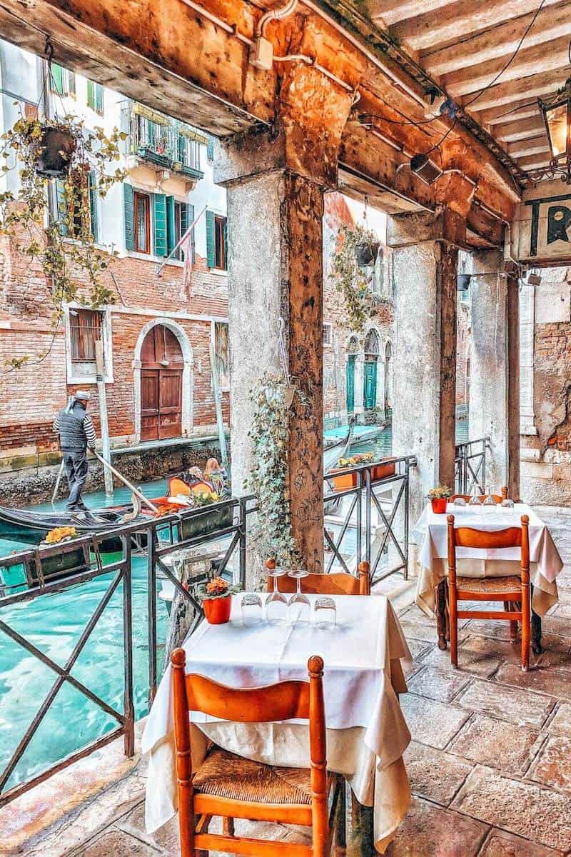 A cafe on a covered terrace next to a canal where a man is standing on a gondola
