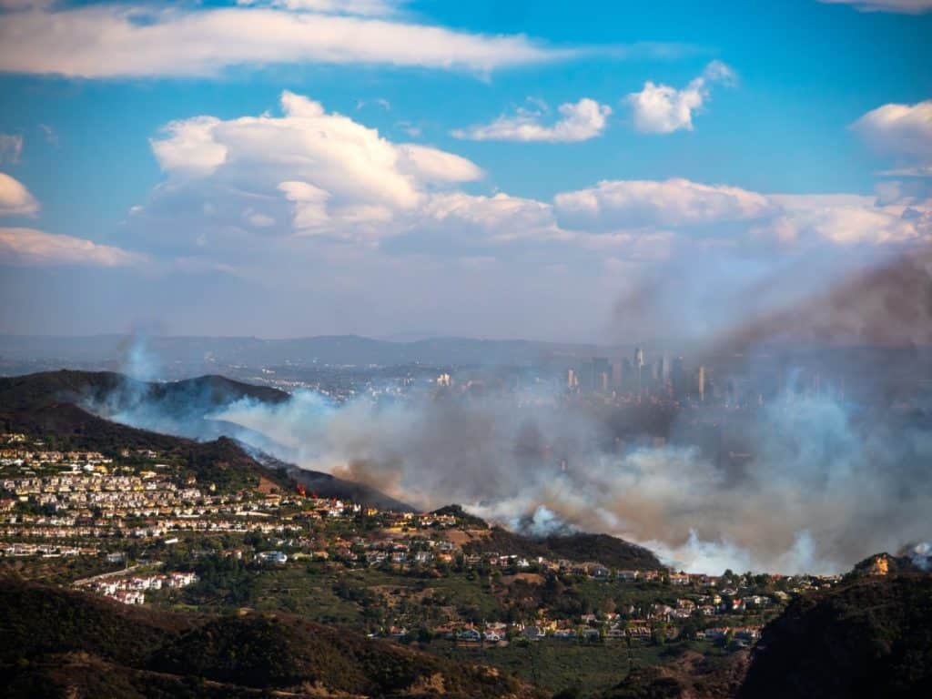 Aerial view of a wildfire spreading through hills near a suburban neighborhood in California, with smoke drifting toward a distant LA skyline under a partly cloudy sky.