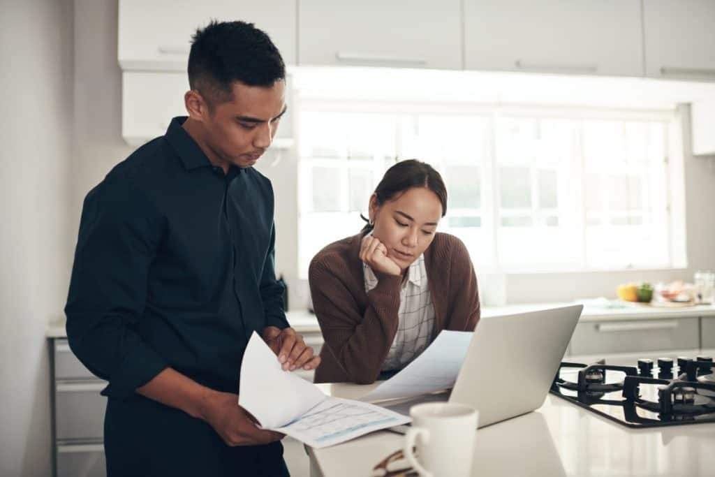 Concerned couple reviewing paperwork and using a laptop in their kitchen after receiving a denial for their fire damage insurance claim.