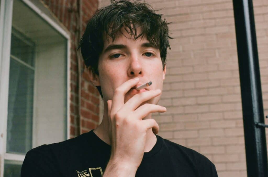 Young man with dark hair smoking a cigarette outdoors, casual attire, brick wall background.