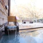 Close-up of an outdoor hot spring bath with wooden deck and a traditional bathing bucket, surrounded by autumn foliage. Private open-air baths like this are a highlight of many Hokkaido onsen towns.