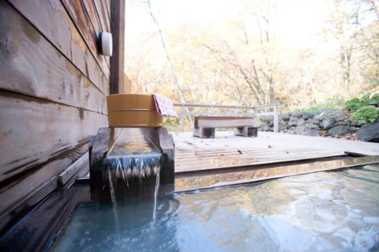 Close-up of an outdoor hot spring bath with wooden deck and a traditional bathing bucket, surrounded by autumn foliage. Private open-air baths like this are a highlight of many Hokkaido onsen towns.