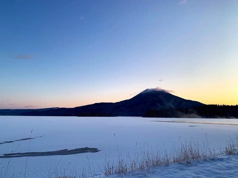 A frozen Lake Akan under a soft sunrise sky with a snow-covered mountain in the distance, viewed from the snowy lakeshore.