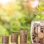 Stacks of coins with growing green plants on top, next to a jar full of change, symbolizing saving money. Budget travel in Hokkaido is possible with smart choices around transport, food, and accommodation.