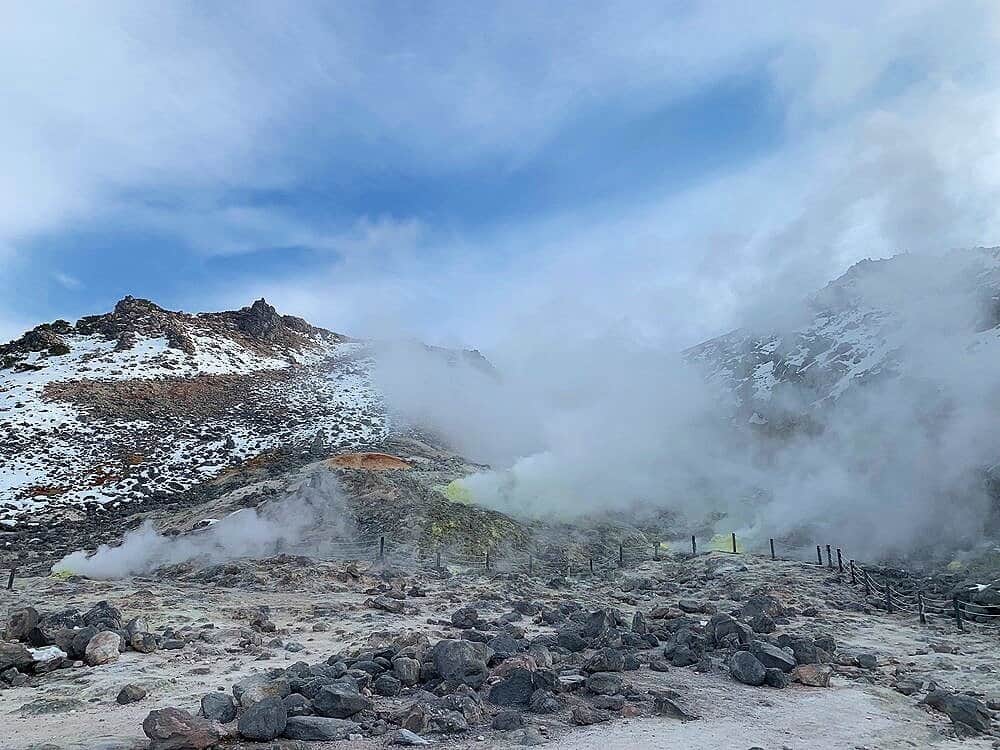 A wide view of Mount Io with white steam rising from scattered sulfur vents, rocky ground, patches of snow, and a partly cloudy blue sky overhead.