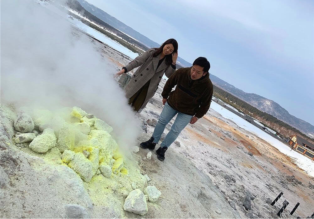 Two people observe steaming sulfur vents surrounded by yellowish sulfur deposits at Mount Io, with snowy ground and mountains in the background.