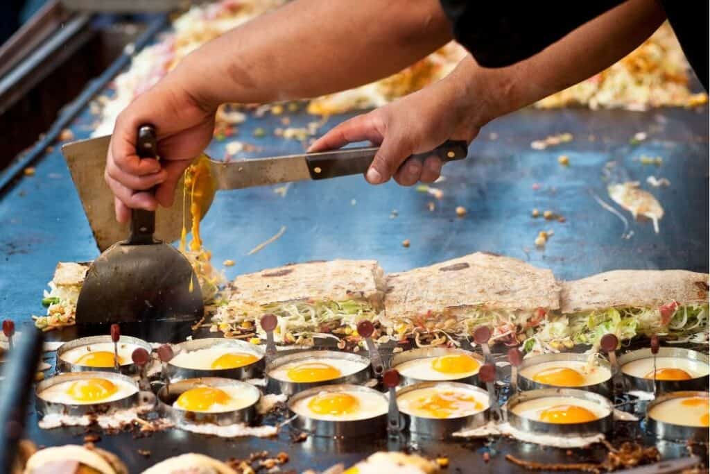 Street food sizzling on a griddle at a Hokkaido summer festival, with a vendor cooking okonomiyaki and sunny-side-up eggs. A vibrant scene that captures the local flavors and lively food stall atmosphere.