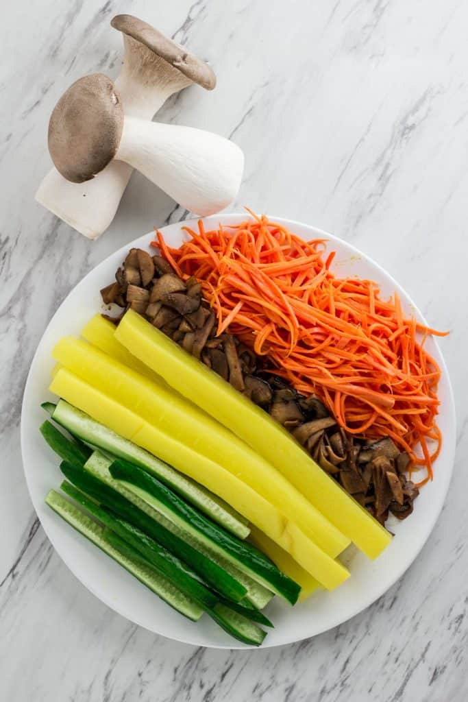Prepared kimbap fillings including bulgogi mushrooms, carrots, cucumber, and Korean pickled radish