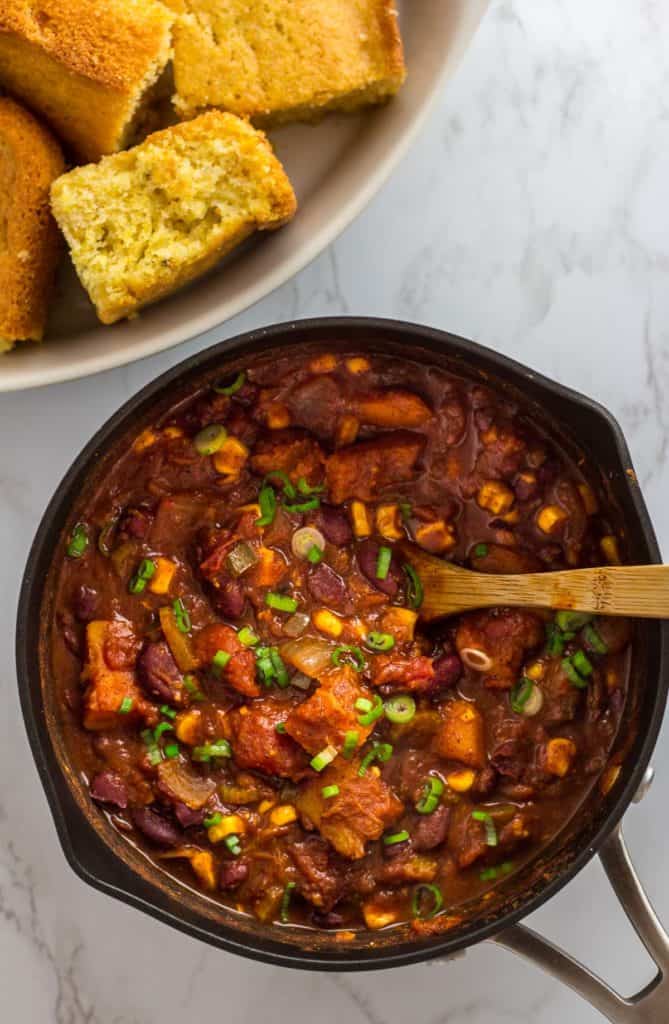Pot of vegan butternut squash chili with beans, corn, and roasted squash, with cornbread in the background.