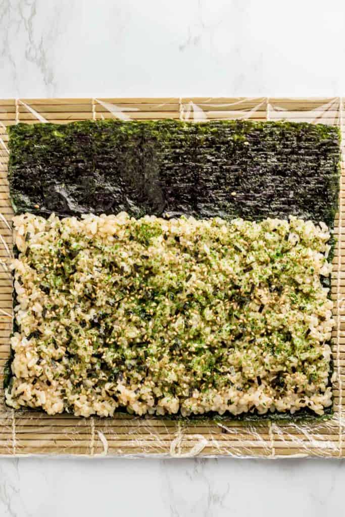 Rice is evenly spread on top of seaweed paper with seaweed flakes sprinkled on top. About one third of the seaweed paper is left empty and not covered with rice.