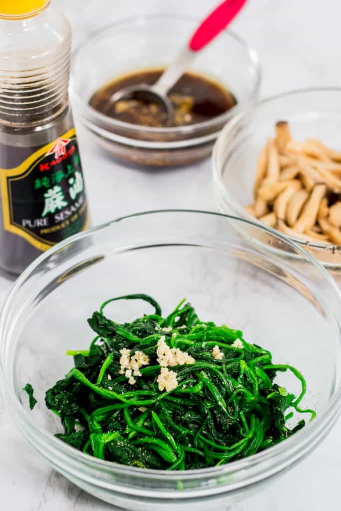 Blanched spinach seasoned with garlic and sesame oil in a glass bowl with soy sauce and mushrooms in the background