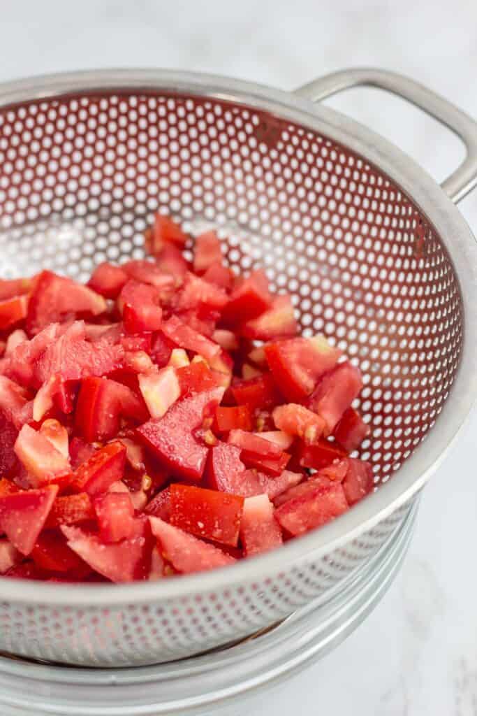 Diced Roma tomatoes are salted and draining in a metal colander.