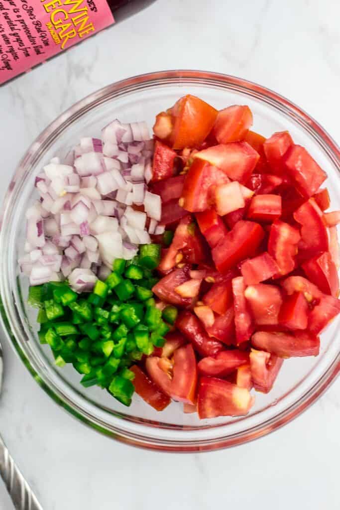 diced roman tomato, finely diced onion and jalapeno in a clear bowl. Red wine vinegar bottle in the background corner.
