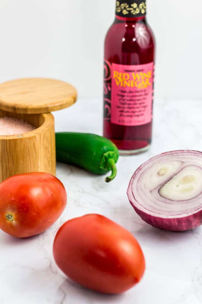 Ingredients for pico de gallo - Roma tomatoes, red onion, jalapeño, salt, and a bottle of red wine vinegar.
