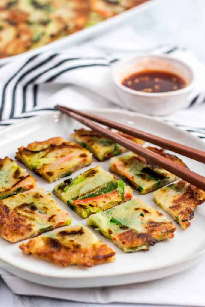 Sliced Korean vegetable pancake served on a plate with chopsticks and dipping sauce on the side.