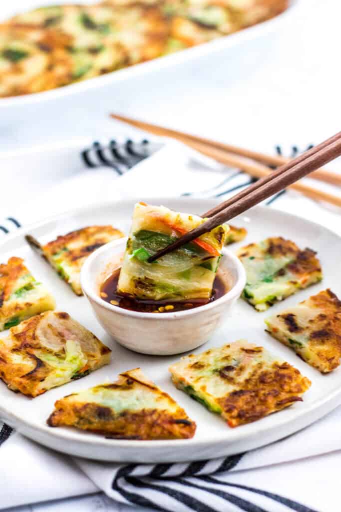 Chopsticks holding a piece of crispy Korean vegetable pancake dipped in soy-based sauce.