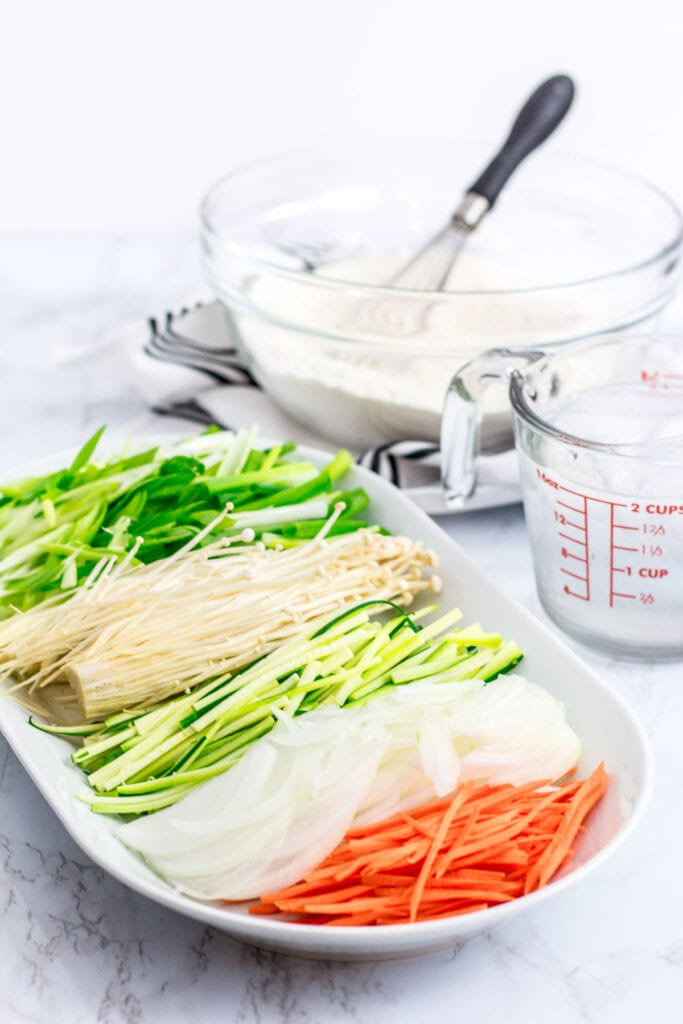 Prep ingredients for Korean vegetable pancake - scallion, onion, carrot, zucchini, and enoki mushrooms on a tray with batter in the background.