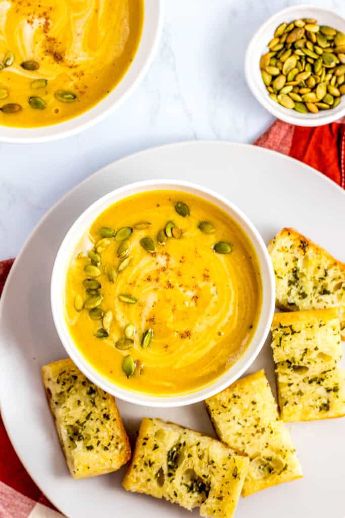 A bowl of creamy vegan butternut squash soup topped with pumpkin seeds, served with garlic bread on the side.