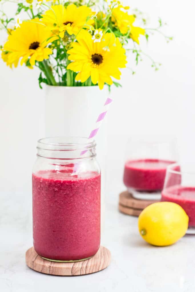 Glass jar filled with vibrant beetroot smoothie with a purple straw, served with lemon and yellow flowers in the background.
