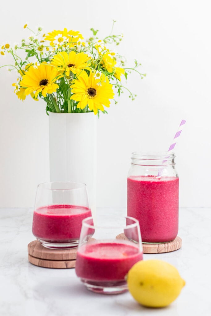 Mason jar and glasses filled with beetroot smoothie next to a vase of yellow flowers and fresh lemon.