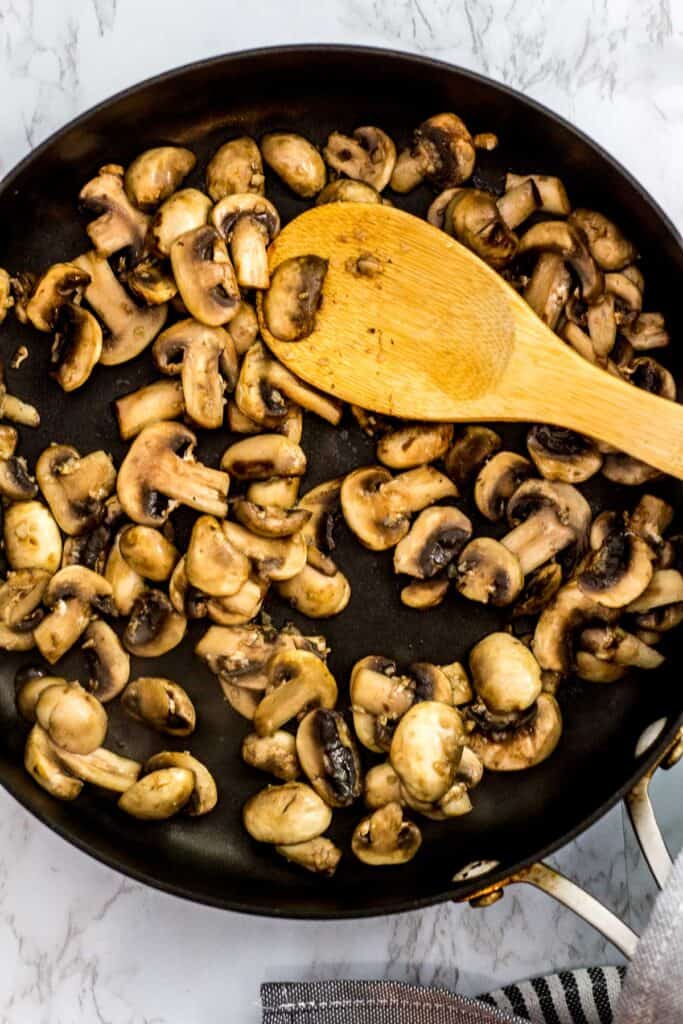 Sliced mushrooms sautéing in a skillet with a wooden spoon.