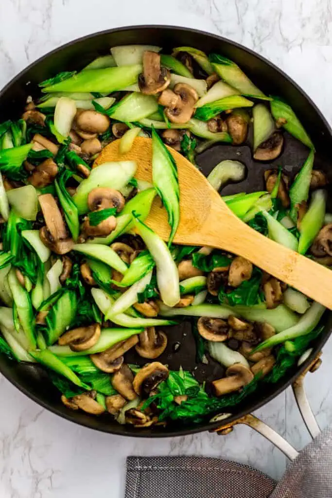 Sliced bok choy stir-fried with mushrooms in a skillet with a wooden spoon.