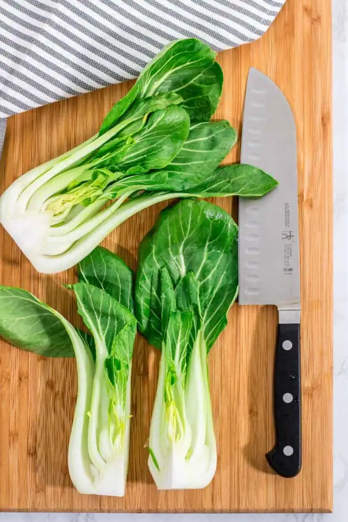 Bok choy sliced lengthwise in half on a cutting board with a chef’s knife.