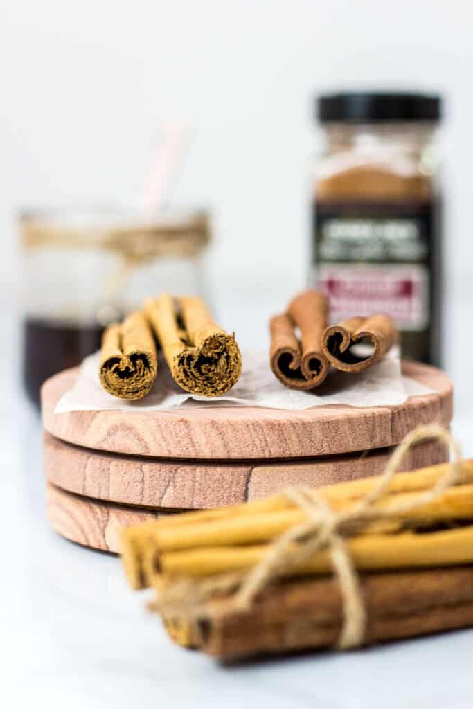 Different types of cinnamon sticks on a wooden board with a jar of homemade cinnamon syrup and ground cinnamon bottle in the background.