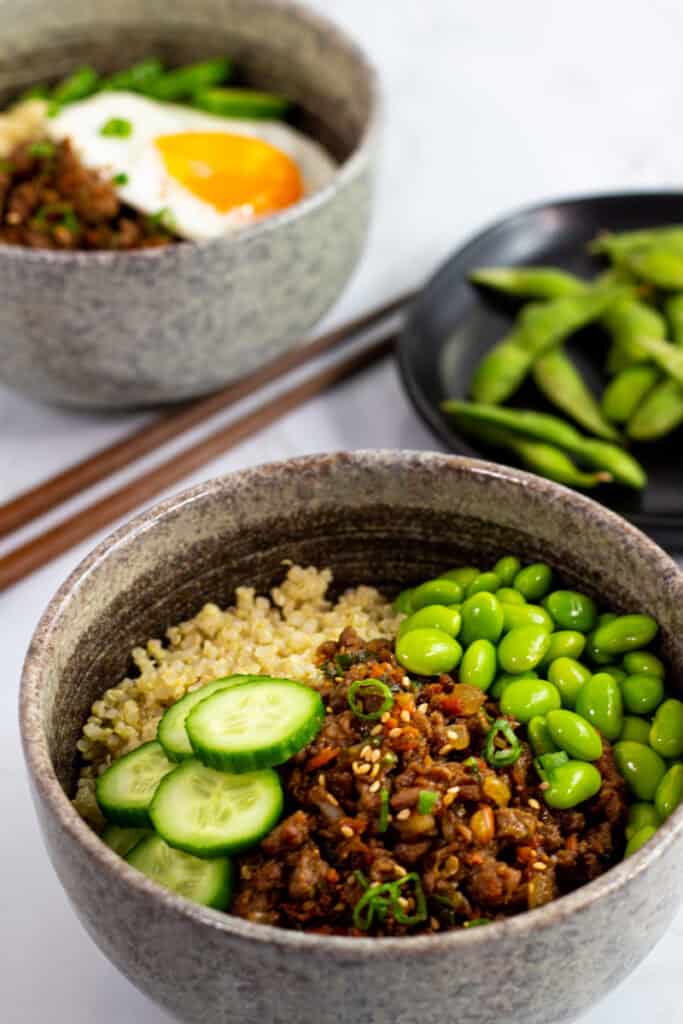 Two bowls of vegan bulgogi, quinoa, cucumbers, and edamame served with chopsticks, one topped with a fried egg for vegetarians