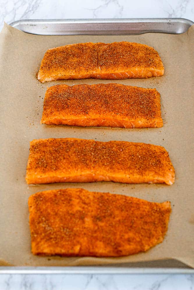 Four salmon fillets on a baking sheet, coated in Old Bay seasoning and spices, read to be baked