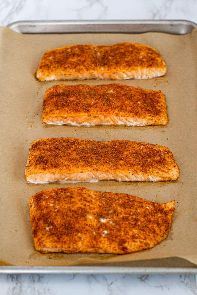 Four baked salmon fillets on parchment paper with golden edges and visible spice rub