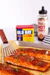 close-up of glazed salmon being lifted with a spatula, with Old Bay and hot honey in the background
