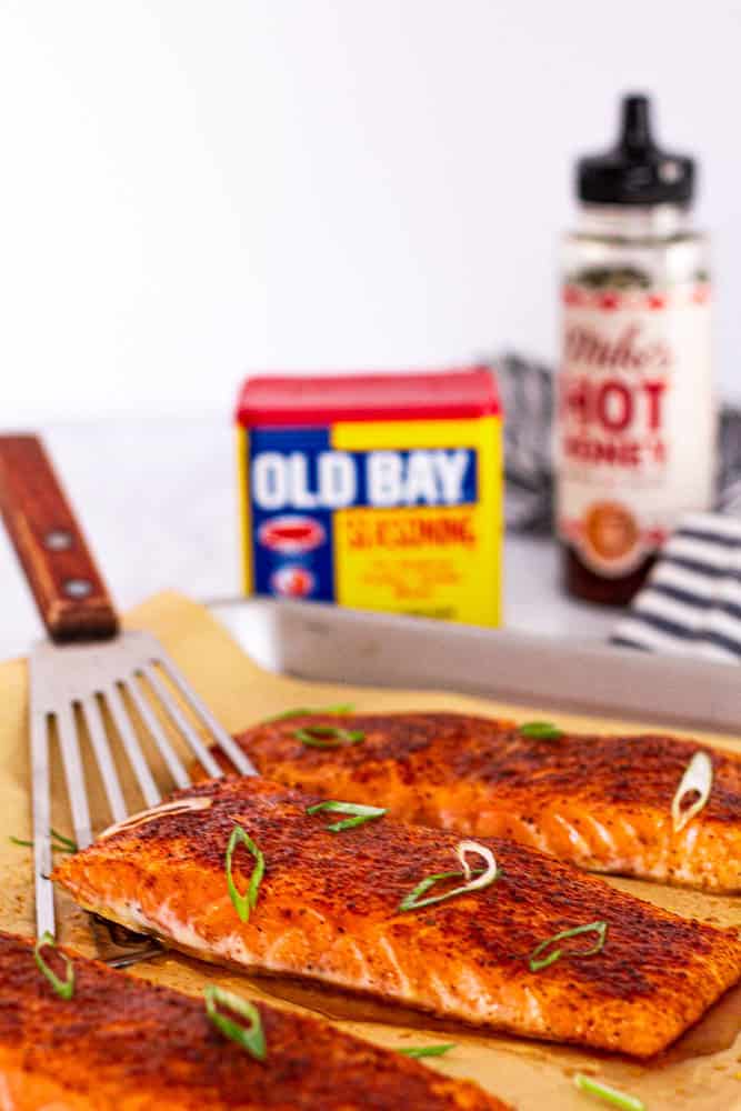 close-up of glazed salmon being lifted with a spatula, with Old Bay and hot honey in the background