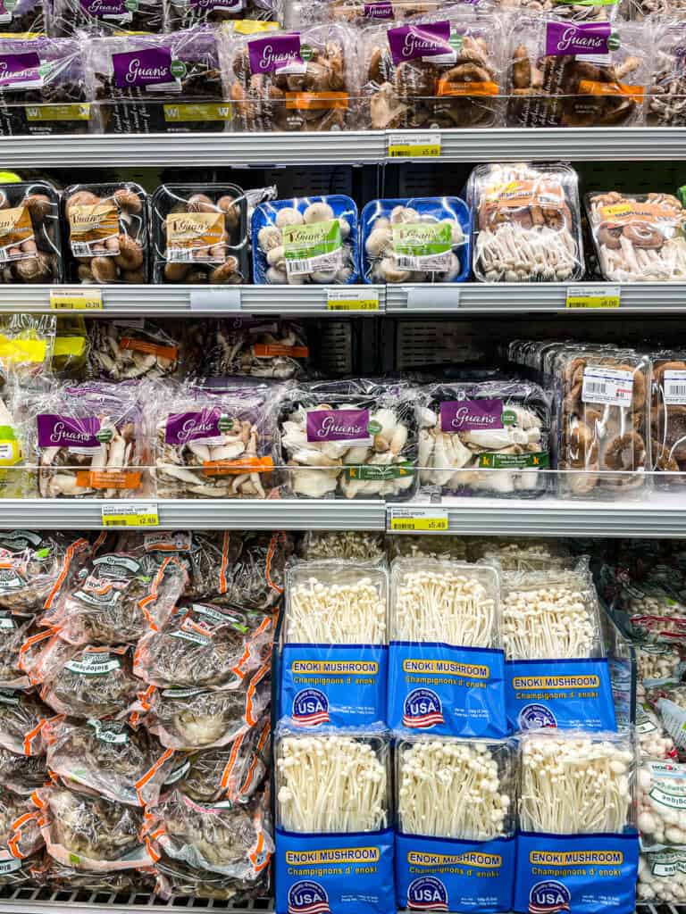 Assorted packages of Japanese mushrooms including enoki, shiitake, and maitake on a grocery store shelf, popular in Japanese soups and hot pots.