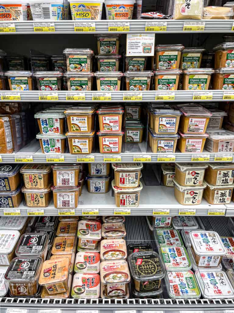 A grocery store shelf stocked with a wide variety of Japanese miso paste containers, including white, red, mellow, organic, and dashi-added miso options.