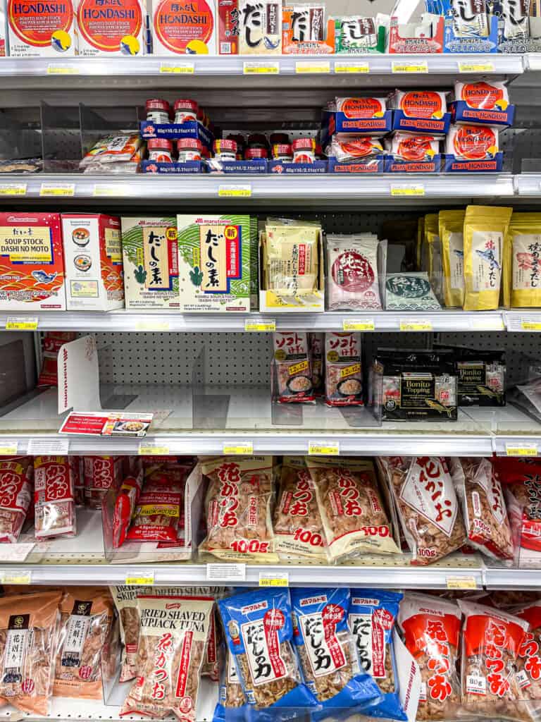 Various packages of dashi soup stock powders and dried bonito flakes (katsuobushi) displayed on a Japanese grocery store shelf.