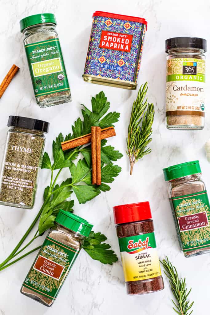 Assorted Mediterranean spices and herbs including oregano, thyme, cumin, smoked paprika, sumac, and fresh parsley on a white background