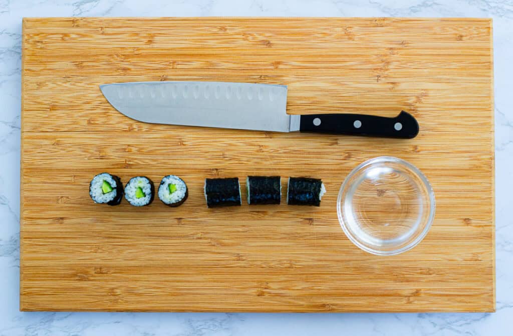 Sliced cucumber maki roll on bamboo cutting board with sharp knife and bowl of water